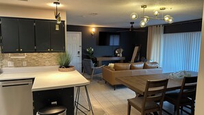 A modern kitchen with dark cabinets, a white countertop, and a built-in dishwasher. There is a dining area with a wooden table and chairs, and a sofa in the background. - The Retreat at Sunset Harbor (Navarre)