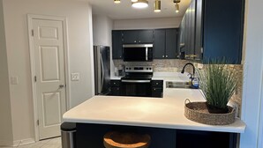 A modern kitchen with dark cabinets, stainless steel appliances, a white countertop, and a hanging light fixture. - The Retreat at Sunset Harbor (Navarre)