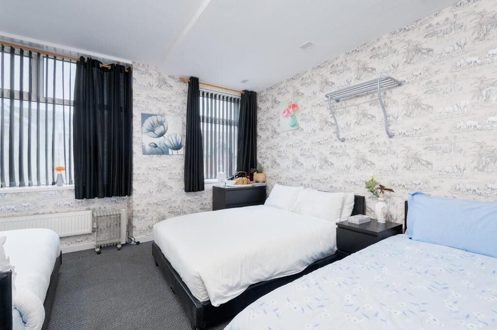 Een hotelkamer met twee bedden, een radiator, een raam met gordijnen, een wandplank met een vaas en een tafeltje met een lamp.