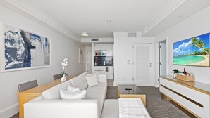 A modern living room with a sofa, coffee table, and a TV on a wall. There is a kitchen in the background.