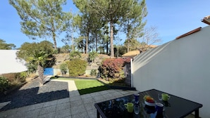 Un patio con una mesa de vidrio, un pequeño jardín con césped y árboles, y una verja azul.