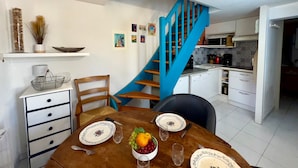 Un comedor con una mesa de madera dispuesta para dos personas, una escalera azul y una cocina al fondo.