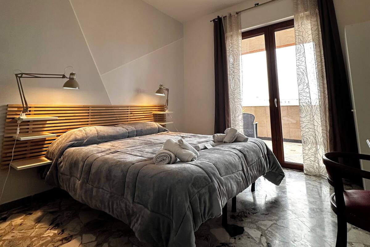 A modern bedroom with a large bed, a bedside table with a lamp, a headboard, and a view of the outside through a window with curtains.