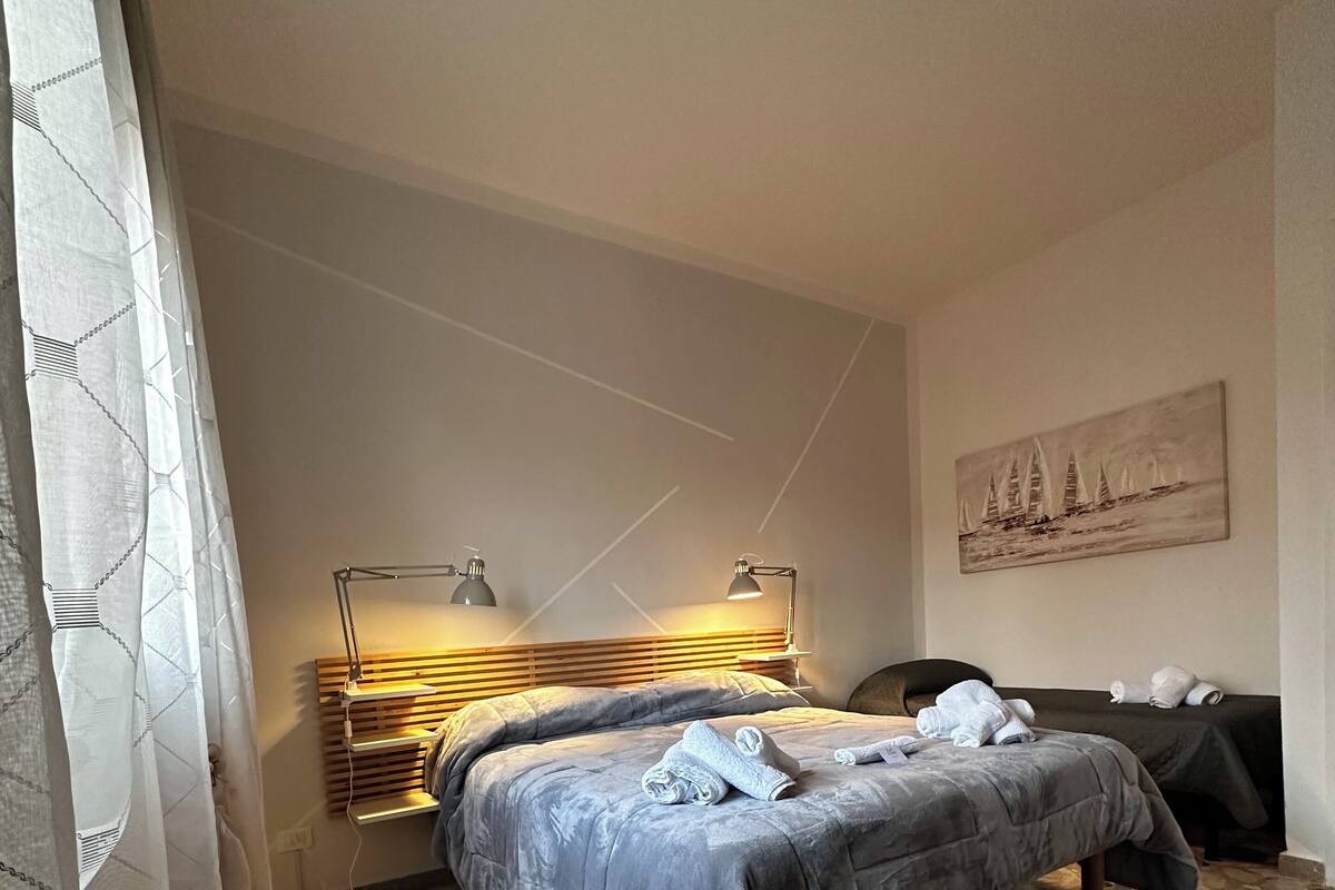 A bedroom with a stone-tiled floor, a bed with a grey duvet, a bedside table with lamps, and a painting on the wall.