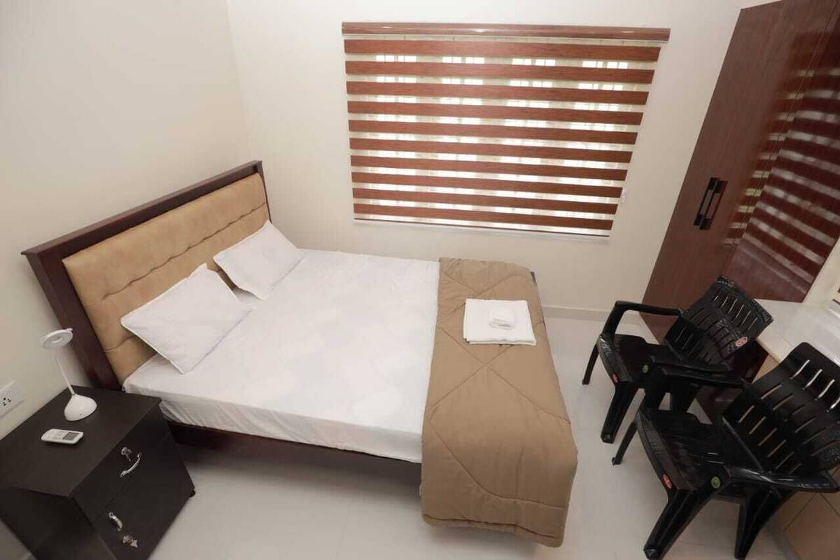 A hotel room with a bed, a bedside table with a lamp, a chair, and a window with blinds.