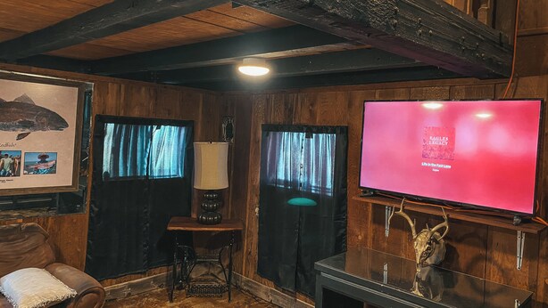 Una habitación rústica con paredes de madera, un televisor montado que muestra 'EAGLES LEGACY', una lámpara y una mesa de madera con una decoración de calavera.