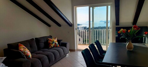 A living room with a dark grey sofa, colorful pillows, a dining table with chairs, and a vase of tulips. A large sliding glass door leads to a balcony with a view of the sea and distant land. - Golden horizon Cavtat - spacious 3-bedroom apartment fully AC (Cavtat)