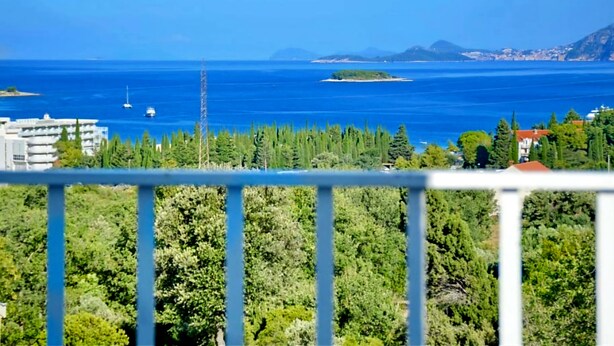 Vista costera con balcón que da a un mar, árboles y edificios a lo lejos.