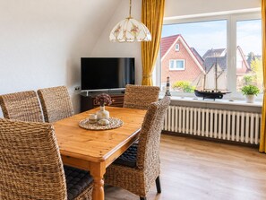 Een eetgedeelte met een houten tafel, rieten stoelen, een televisie en uitzicht op huizen door een raam.