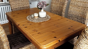 Een eetgedeelte met een houten tafel, rieten stoelen, een vaas met bloemen en een wandlamp.