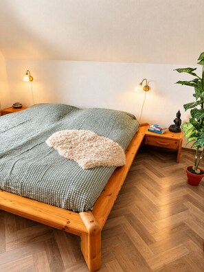 Een bed met een groen geweven sprei, een zacht kussen en een houten hoofdbord. Twee nachtkastjes met lampen en een potplant.
