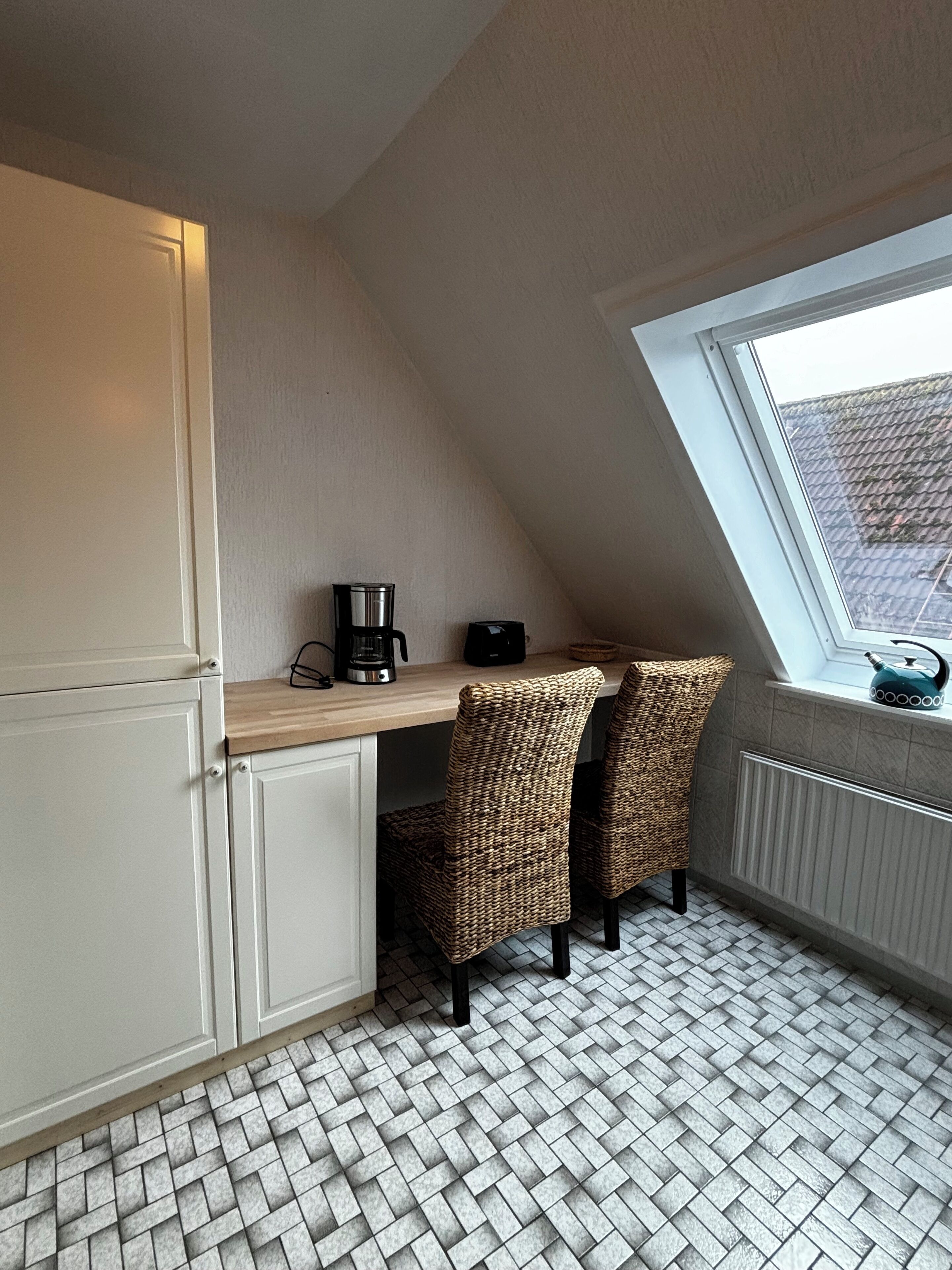 A small kitchen with a wooden countertop, two wicker chairs, a coffee maker, and a window with a view of a roofed area.