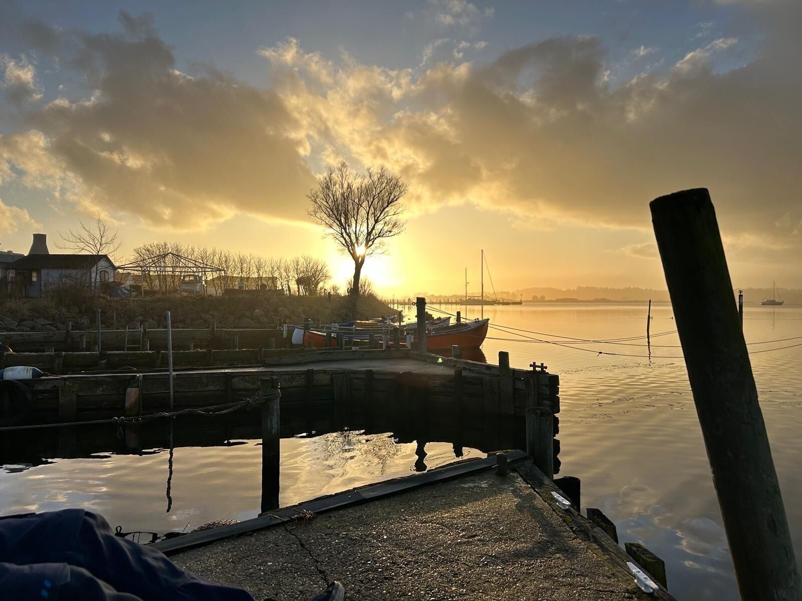 Un muelle con barcos amarrados, una masa de agua tranquila y una puesta de sol al fondo.