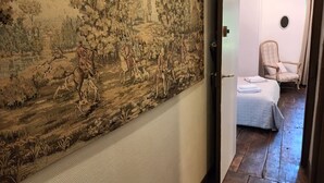 Ein Korridor mit großem Wandteppich, der eine historische Szene zeigt, Holzboden, Bett mit weißer Bettdecke und Stuhl mit Karomuster.