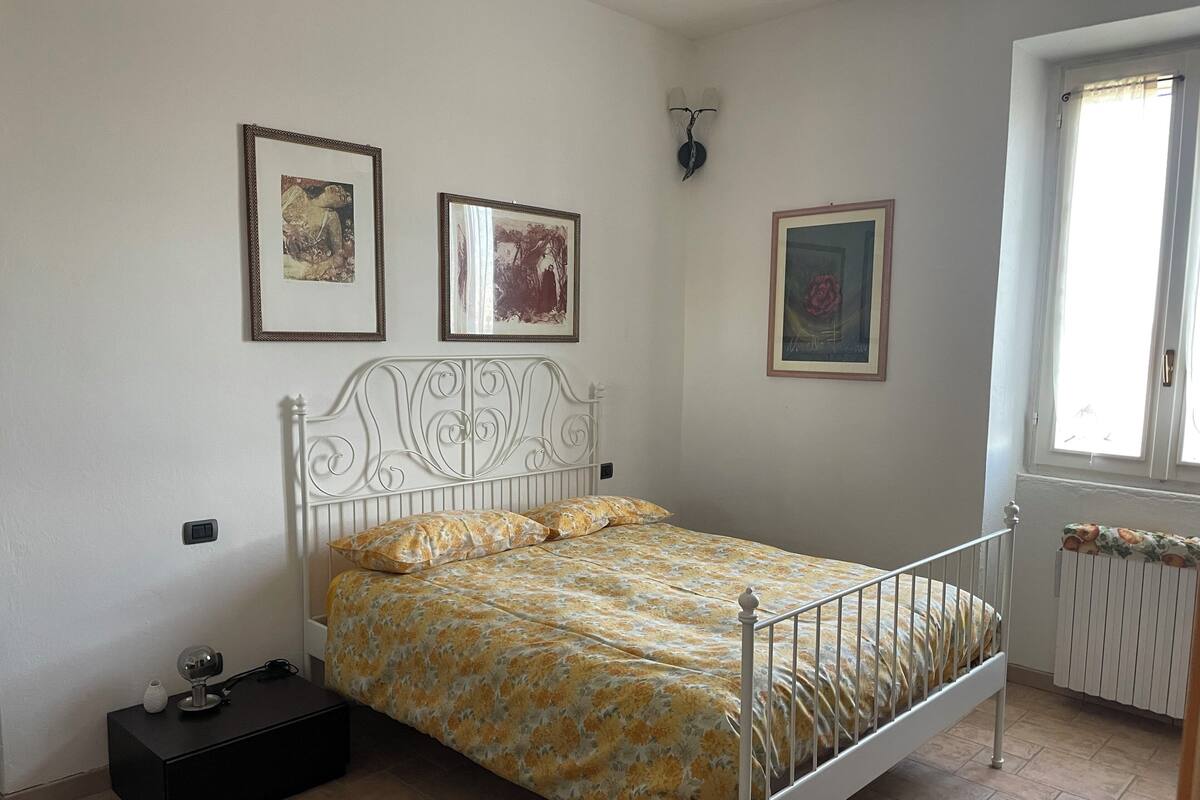 Een slaapkamer met een bed, een nachtkastje, een wandlamp en kunstwerken in lijsten.