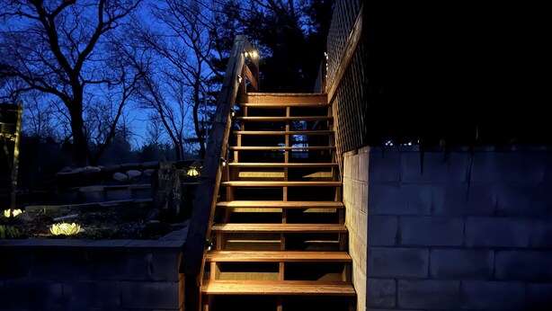 A wooden staircase leading up to a building, surrounded by a stone wall and a garden with a stone bench and a lamp post.