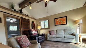 A living room with a leather sofa, a black leather chair, a beige couch, wooden flooring, a ceiling fan, and a large window with a view of trees.