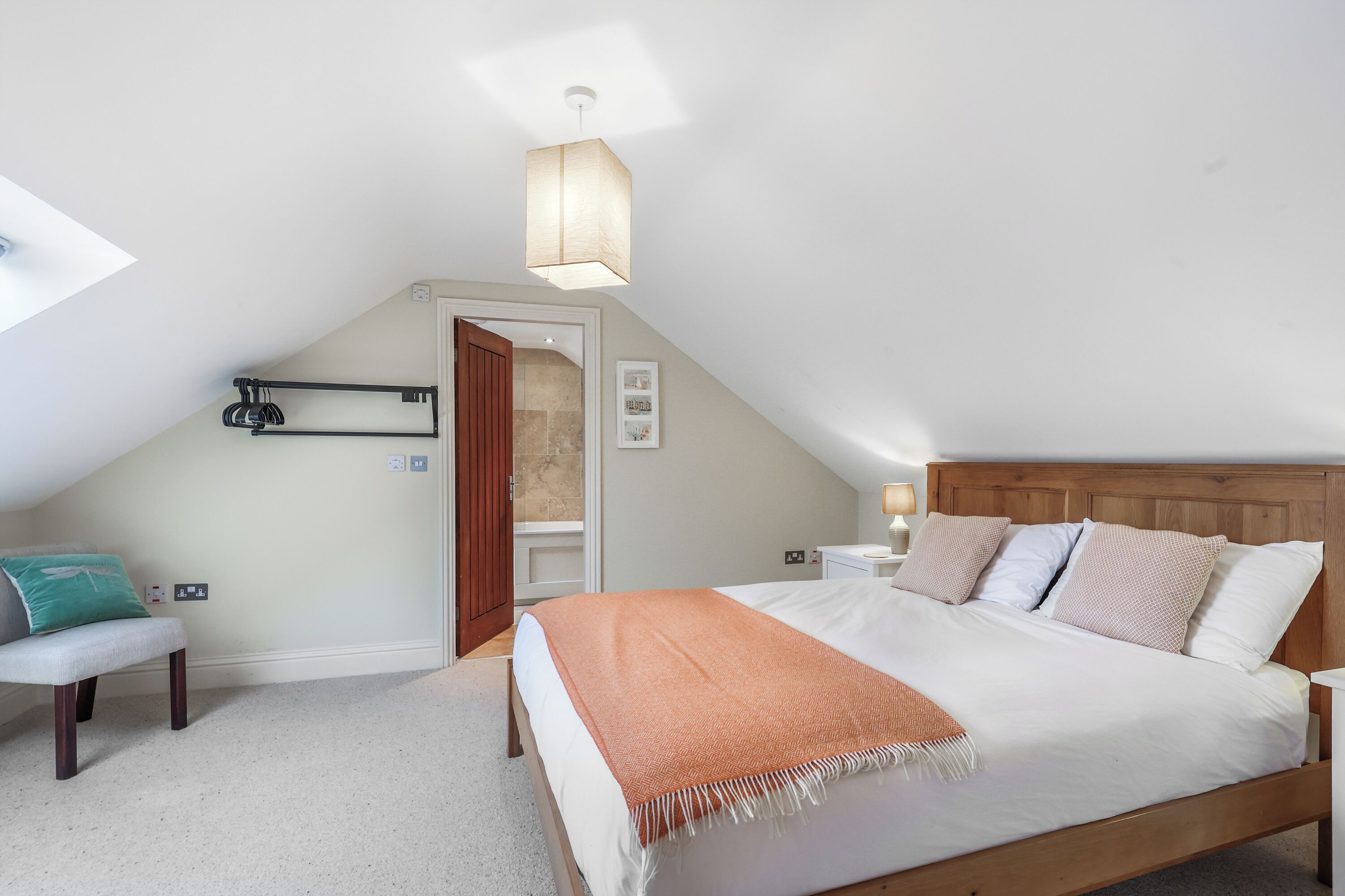 A bedroom with a large bed, a wooden headboard, a bench, a hanging light fixture, and a door leading to a bathroom.