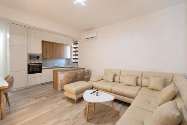 A modern living room with a beige sofa, a small white table, and a kitchen area with white cabinets and a microwave.