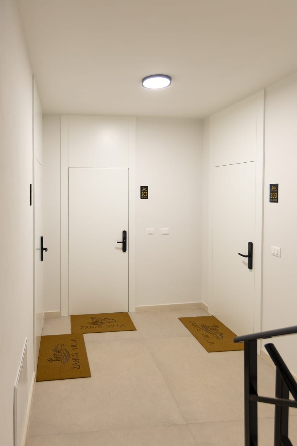 A hallway with white doors, each marked with a number. There are two brown doormats on the floor.