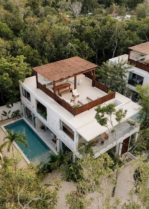 A modern multi-story house with a rooftop patio, a swimming pool, and lush surrounding vegetation.