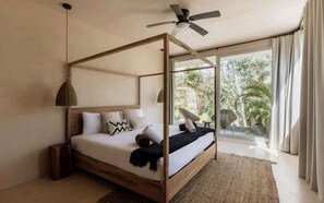 A bedroom with a wooden four-poster bed, a ceiling fan, and a view of greenery outside.