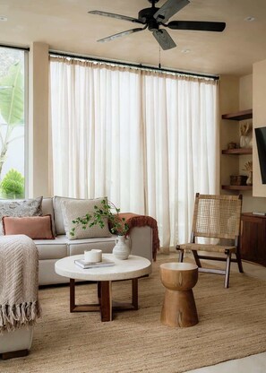 Un salon comprenant un canapĂ©, une table basse ronde, une table dâappoint en bois, un fauteuil en osier et un ventilateur de plafond.
