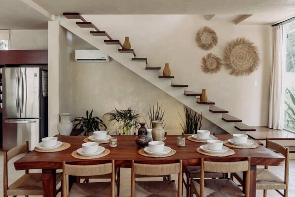 Un coin repas avec une table en bois dressĂ©e pour six personnes, un escalier ornĂ© de vases dĂ©coratifs et une cuisine Ă©quipĂ©e dâun rĂ©frigĂ©rateur en acier inoxydable.