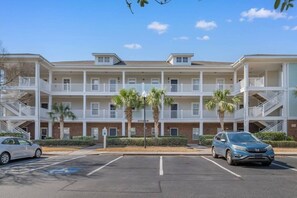 A multi-story building with a parking lot in front, featuring a central entrance and multiple balconies. - Stylish Barefoot Golf Condo with Pool Access (North Myrtle Beach)