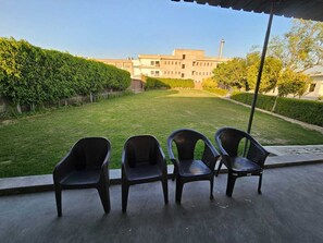 A covered outdoor seating area with four chairs, a well-maintained lawn, and a building in the background. - Govind Resort (Mathura)