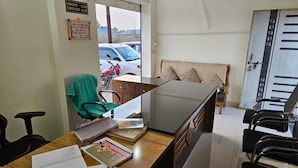 A room with a desk cluttered with papers, a calculator, and a green chair. A sofa with patterned cushions is against the wall, and a door is visible in the background. - Govind Resort (Mathura)