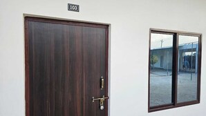 A dark wooden door with a brass handle and lock, a small window with a view of a building and a person outside, and a number sign above the door. - Govind Resort (Mathura)