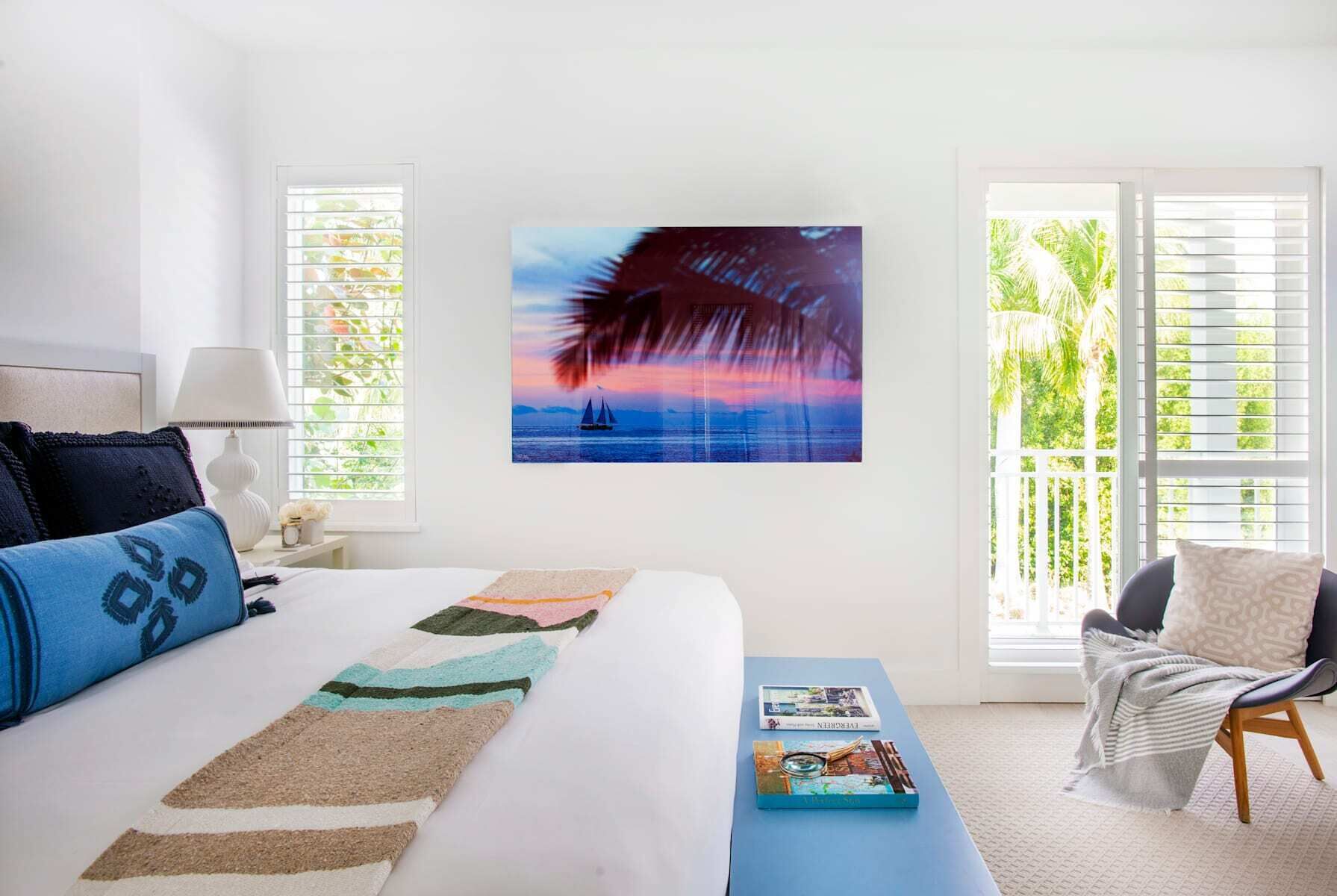 A bedroom with a bed, a bedside table, a chair, a window with blinds, and a painting of a sunset over water.