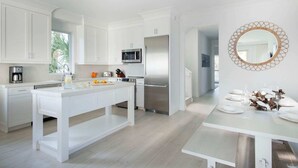 Una cocina moderna con armarios blancos, una isla central, electrodomésticos de acero inoxidable y un comedor con una mesa y sillas blancas.