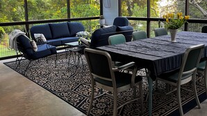 A sunlit room with a blue sofa, dining table, and chairs, large windows offering a view of greenery. - Historic 3-bedroom home in beautiful Athens. Walkable to town and Campus. (Athens)