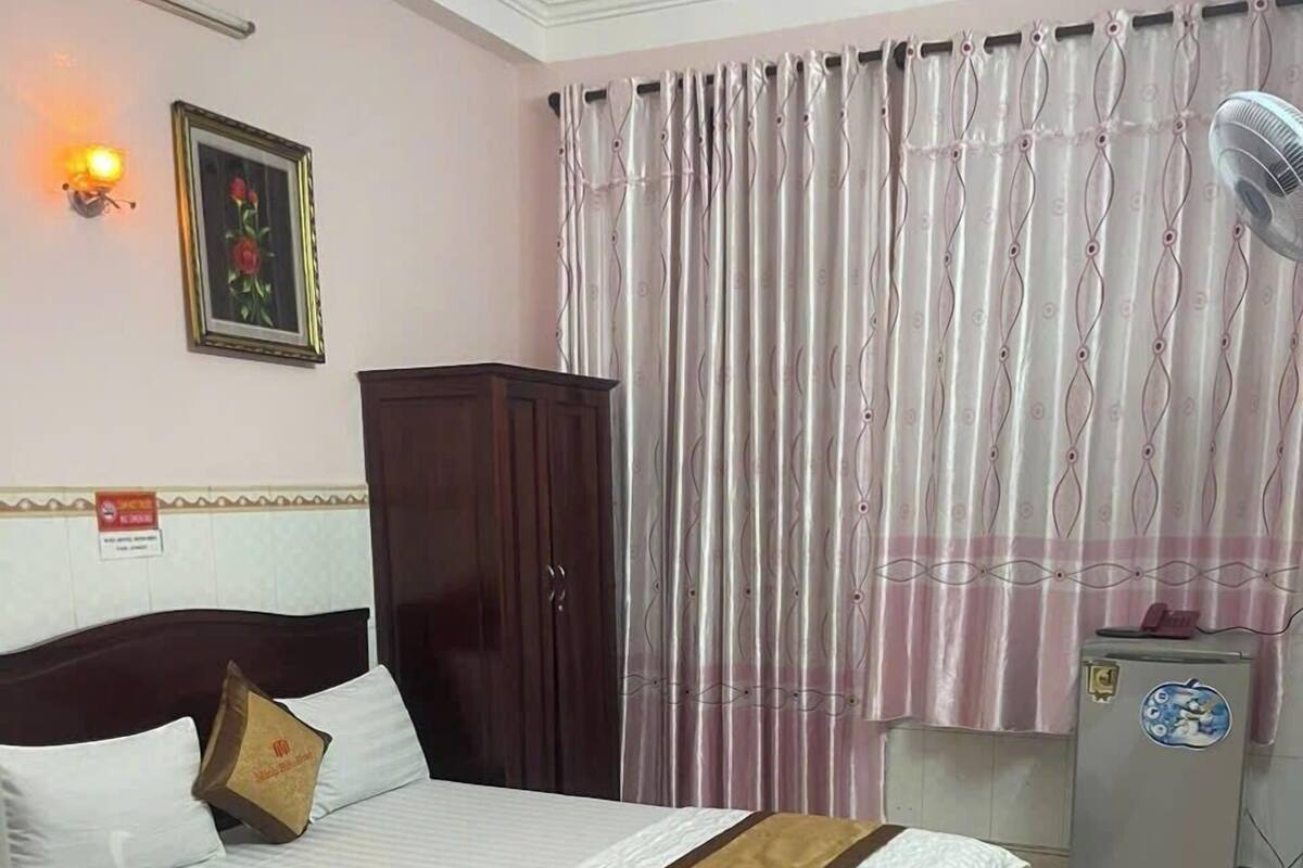 Une chambre d’hôtel comprenant un lit, une armoire, un bureau, une chaise, une table de chevet, un ventilateur et une fenêtre avec des rideaux.