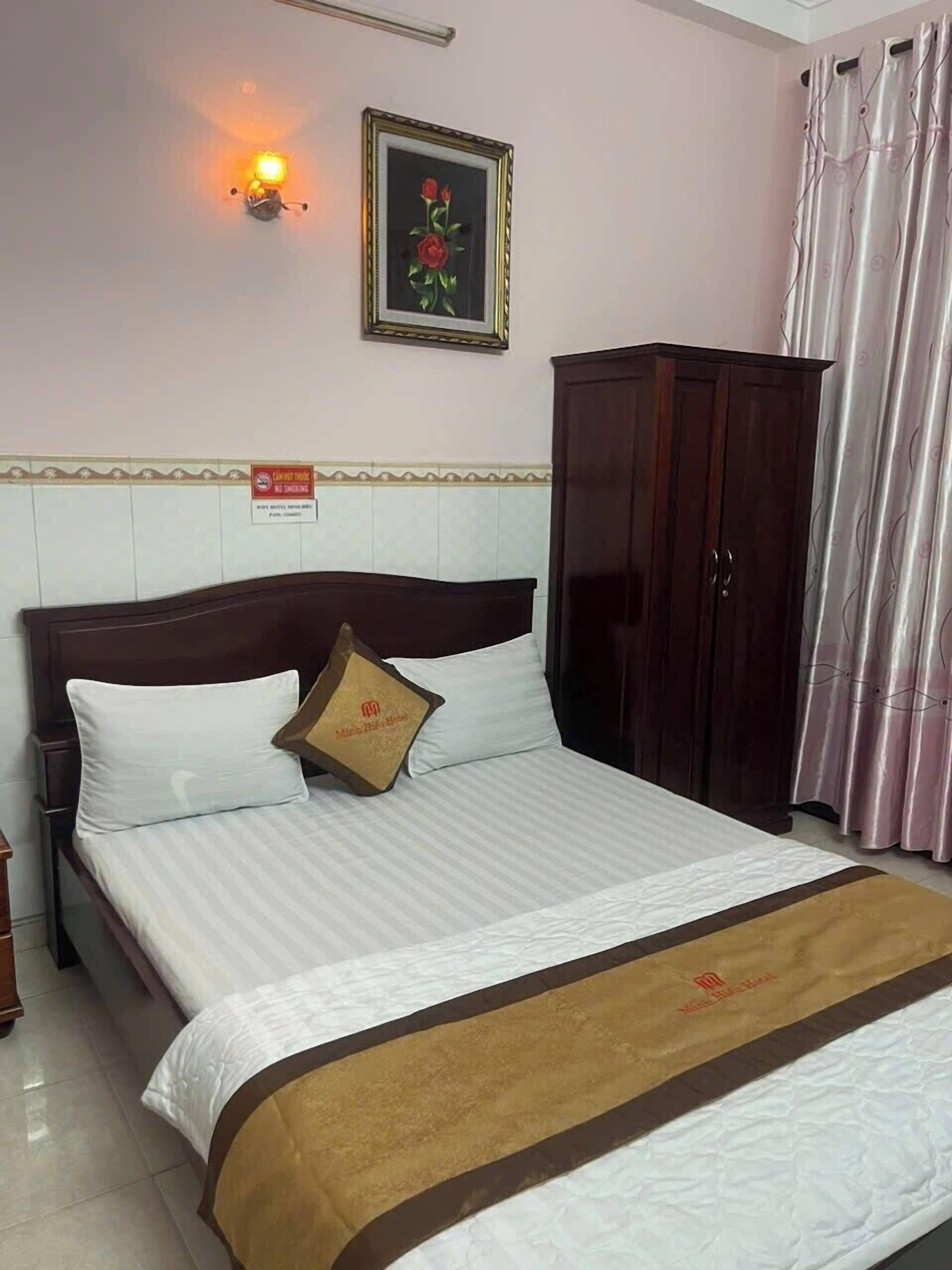 A neatly made bed with a white and brown bedspread, a wooden headboard, and a single decorative pillow. A framed picture hangs on the wall above the bed, and a dark wooden wardrobe is to the right.