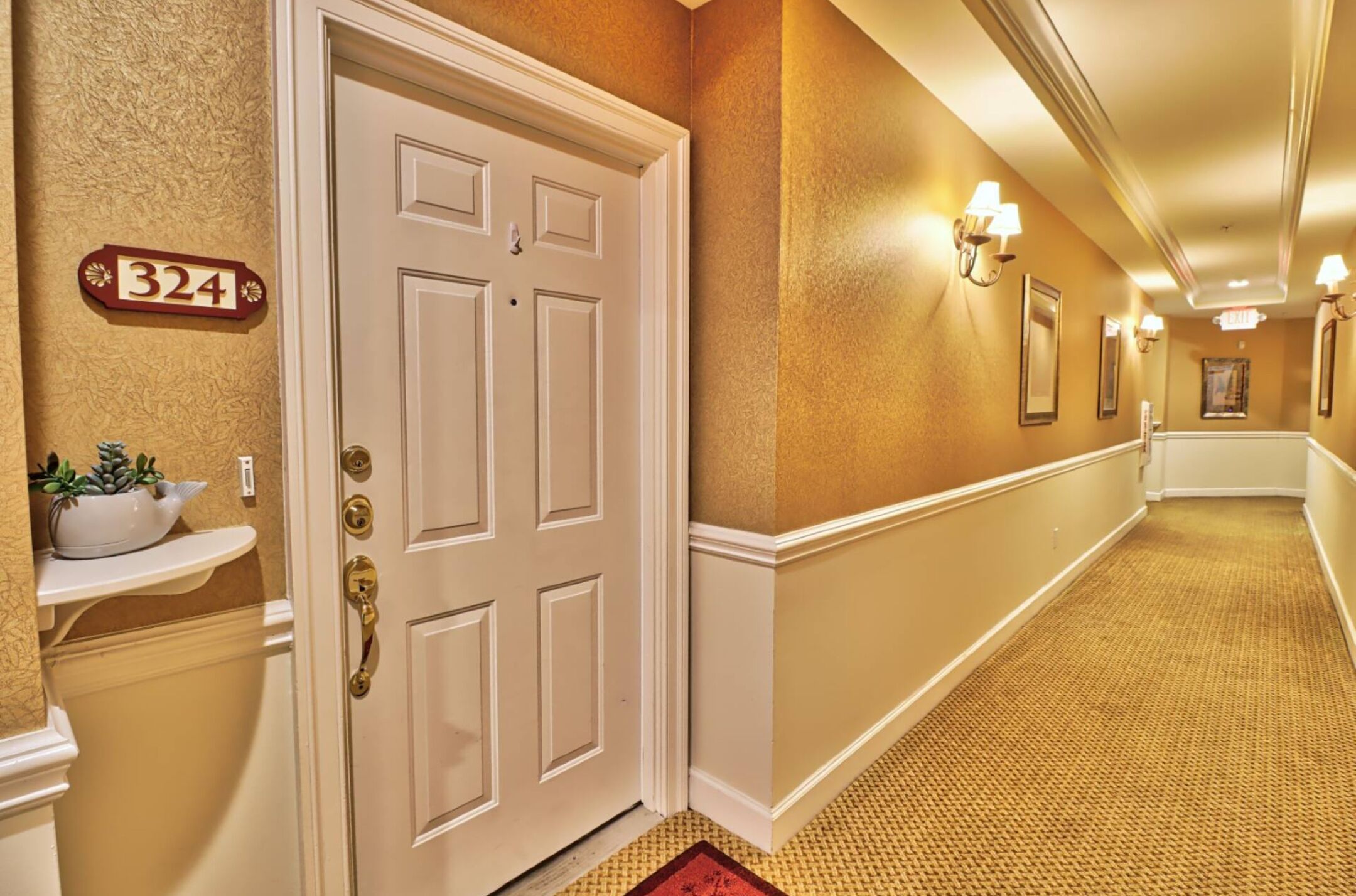 A hotel hallway with a white door, a number sign, a plant, and wall sconces.