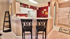 A kitchen with a white countertop, two black chairs, a refrigerator, and a red wall with artwork. - Beautiful Barefoot Golf Resort Condo (North Myrtle Beach)