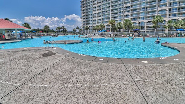 A large outdoor swimming pool surrounded by lounge chairs and umbrellas, with a multi-story hotel in the background. - Beautiful Barefoot Golf Resort Condo (North Myrtle Beach)