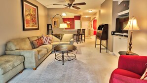 A living room with a sofa, a coffee table, a red chair, a ceiling fan, and a kitchen area in the background. - Beautiful Barefoot Golf Resort Condo (North Myrtle Beach)