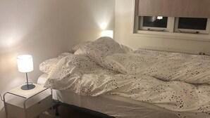A bedroom with a bed, a bedside table with a lamp, a window with blinds, and a rug. - Holidayhome De Molen (Noordwijk)