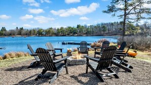 A lakeside seating area with Adirondack chairs around a fire pit, overlooking a calm lake with boats and docked canoes. - Lakefront Summer Getaway w/ HotTub+Games+FirePit (Long Pond)