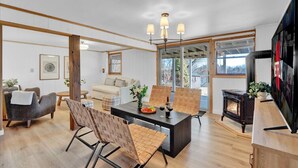 A modern living room with a wooden floor, a dining table with chairs, a sofa, a fireplace, and a television. - Lakefront Summer Getaway w/ HotTub+Games+FirePit (Long Pond)