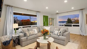A modern living room with a grey sectional sofa, a round coffee table, and a view of a sunset over a body of water. - Luxury Spa, Patio, Deck & Views of Puget Sound in the Heart of Old Town, Tacoma (Tacoma)