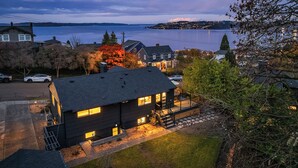 A modern house with a dark roof, a well-lit front porch, and a view of a lake and distant hills. - Luxury Spa, Patio, Deck & Views of Puget Sound in the Heart of Old Town, Tacoma (Tacoma)