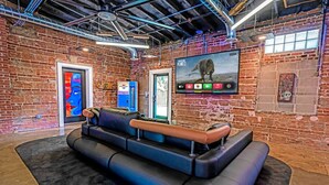 A modern lounge area with a black leather couch, a large flat-screen TV mounted on the wall, and a brick wall with exposed ceiling pipes. - b3 Room4: Begin King Suite (Sparta)