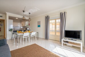 A modern living room with a flat-screen TV, a dining area with a table and chairs, and a kitchen in the background.
