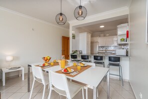 A modern dining area with a white table and chairs, a breakfast setting with fruits and juice, and a kitchen in the background.
