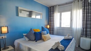 A bedroom with a bed, pillows, a bedside table, a lamp, a window, and curtains. - Chic Sea View Apartment by Hello Homes Sitges (Sitges)